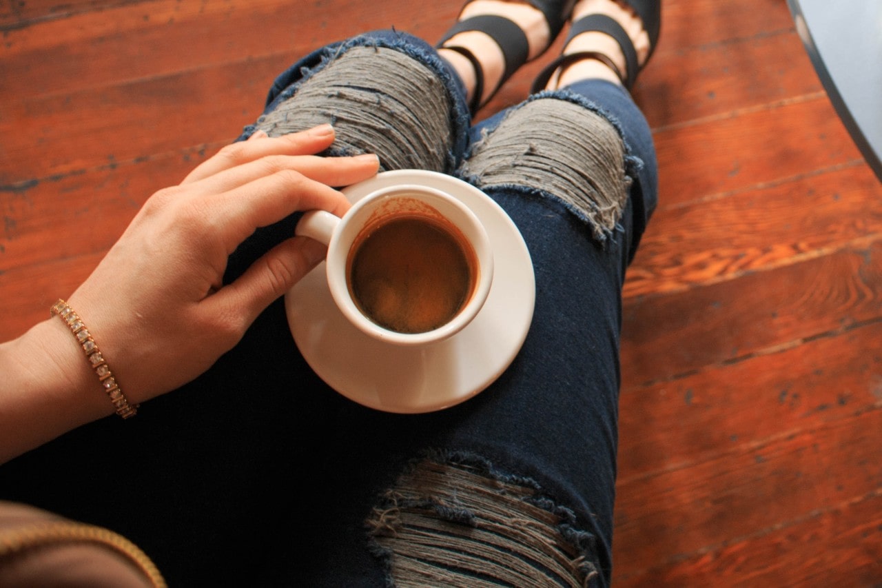 A woman sitting on the floor sipping coffee wears a gold tennis bracelet.