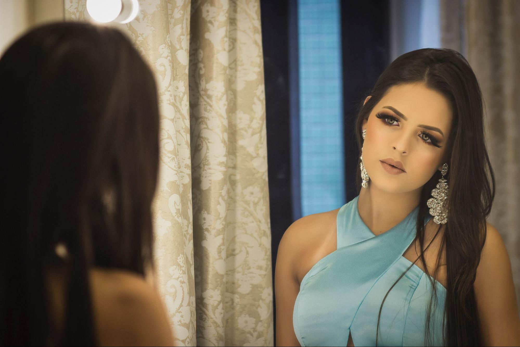 a woman in a blue formal dress and wearing dramatic diamond earrings looks in the mirror.