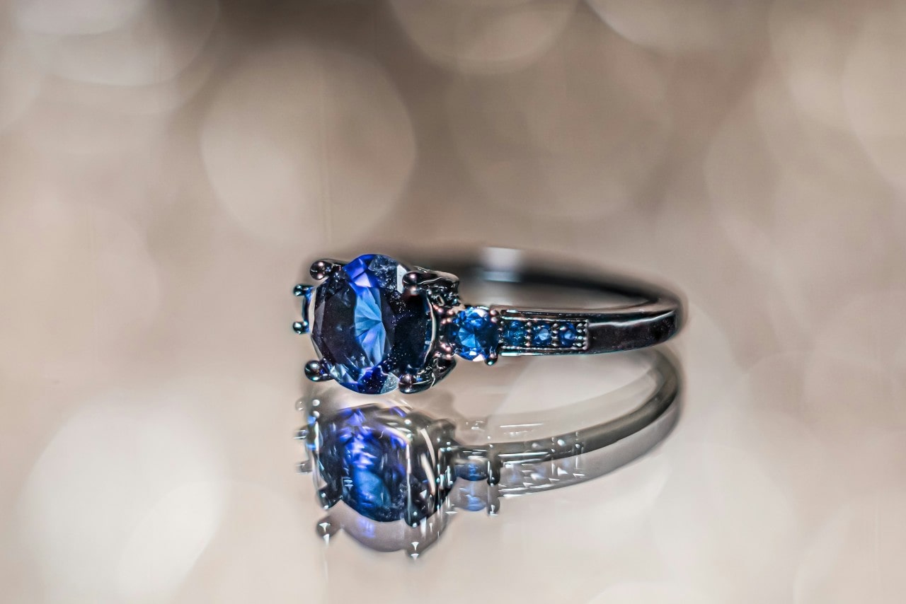 A close up of a white gold engagement ring with a sapphire center stone and smaller sapphire accents against a blurred background.