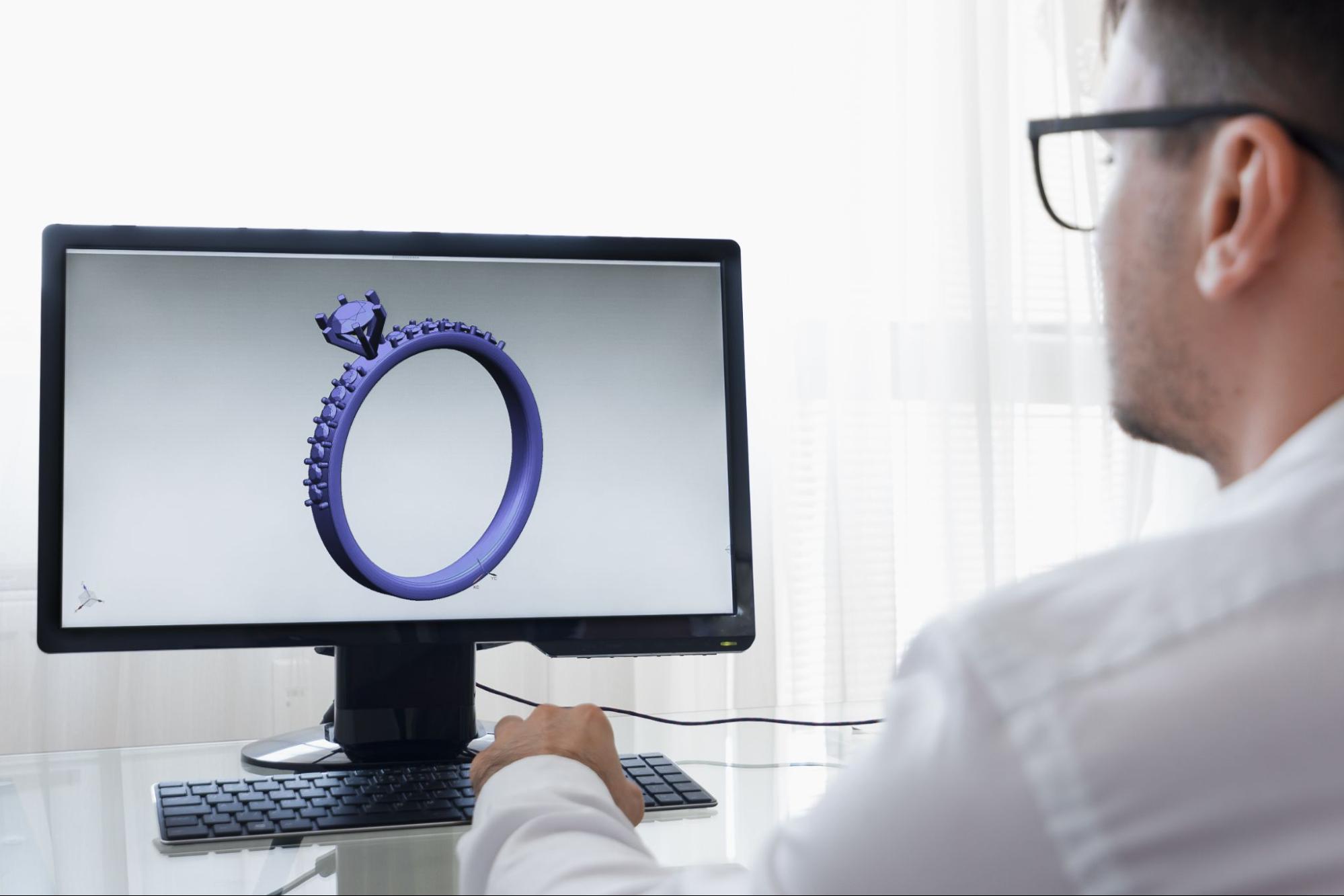 A jeweler using CAD software for jewelry design views a 3D digital model of a diamond engagement ring on a computer screen.