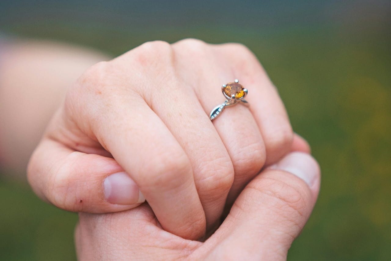 A cushion cut citrine on a white gold entwined band with diamond accents on the hand of a woman holding the hand of her spouse-to-be.