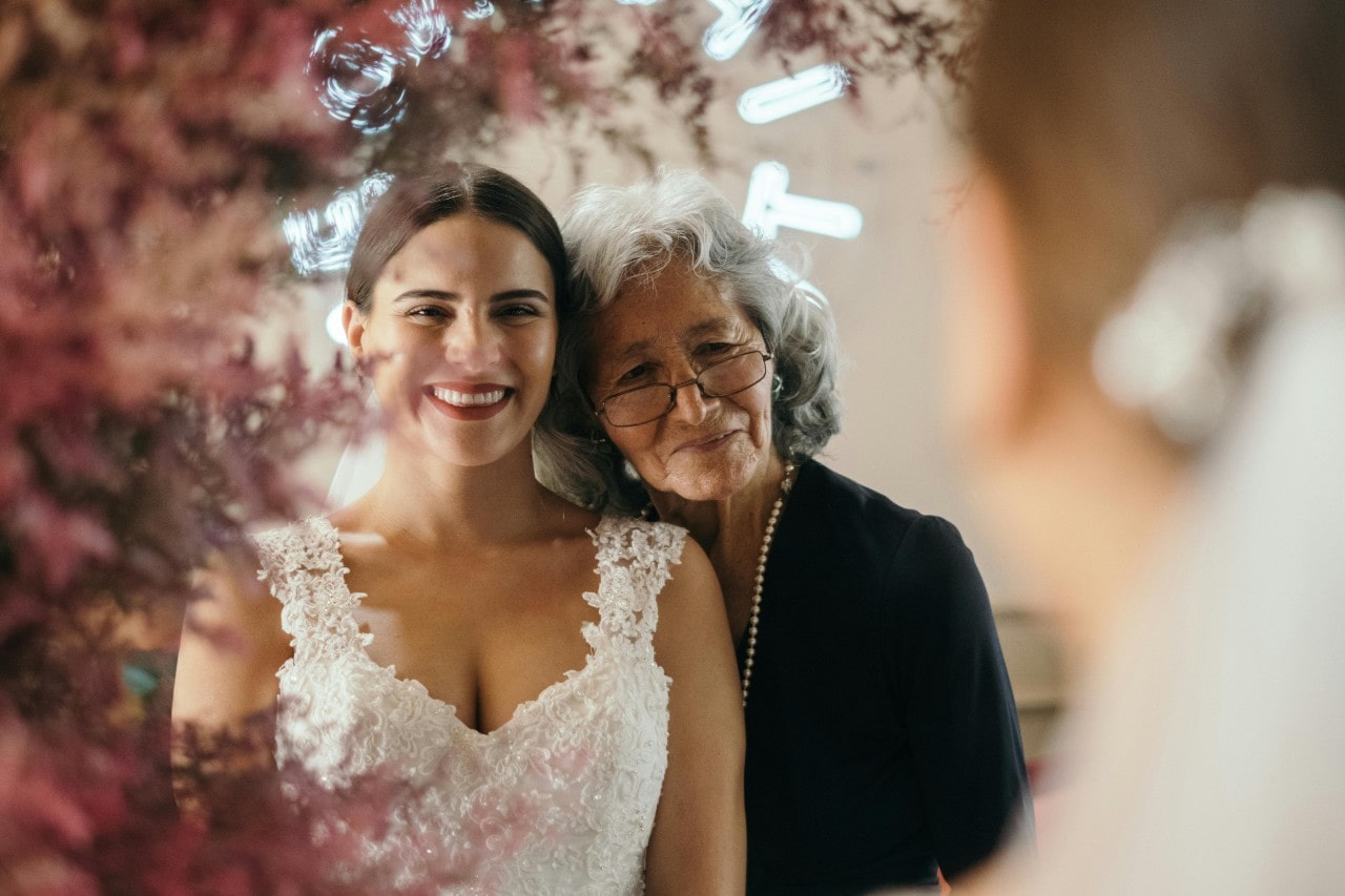 A bride in a white lace dress smiles warmly while standing next to an older woman wearing glasses and black attire, framed by pink flowers.