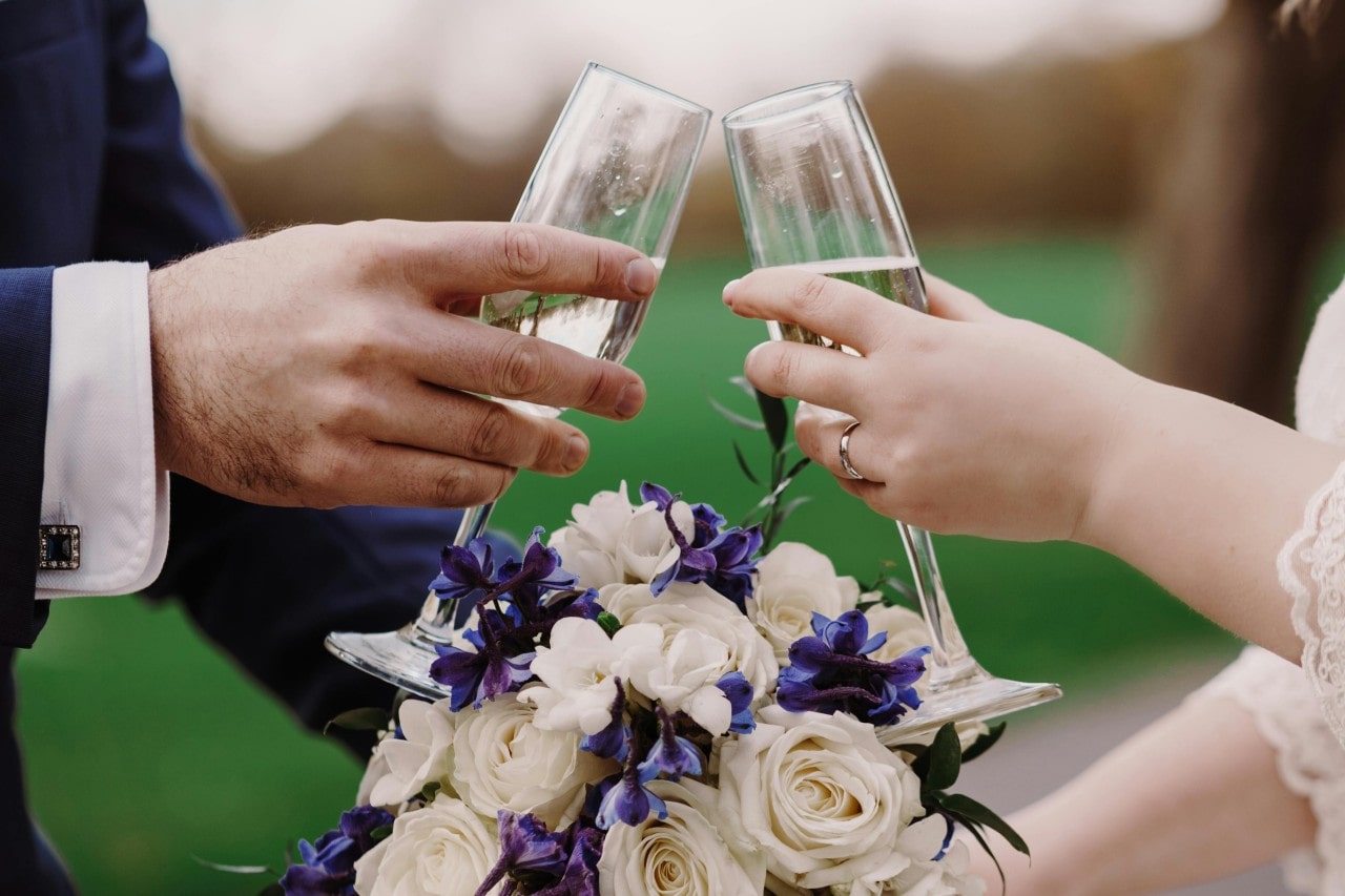 Close up of the bride and groom’s hands clinking champagne glasses over a bouquet of white and purple flowers.