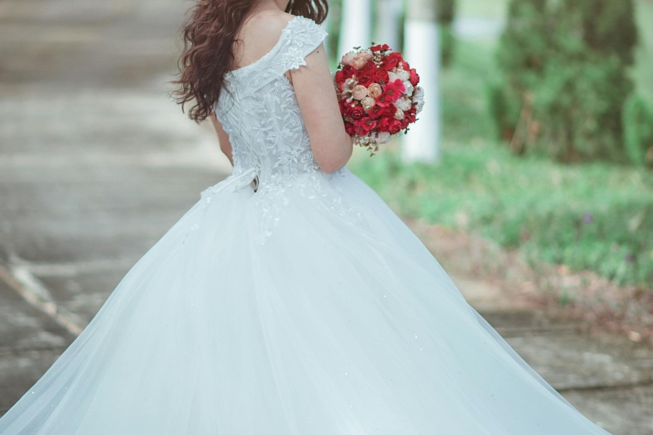 A bride in a flowing white gown holds a vibrant bouquet of red and peach flowers, walking along a garden path.