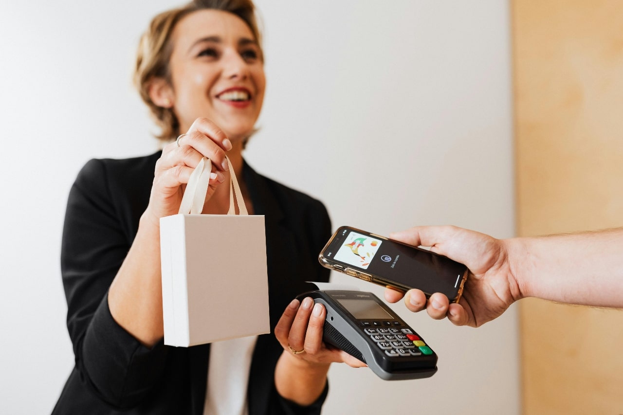 A smiling woman hands a shopping bag to a customer making a contactless payment with a smartphone over a card reader.