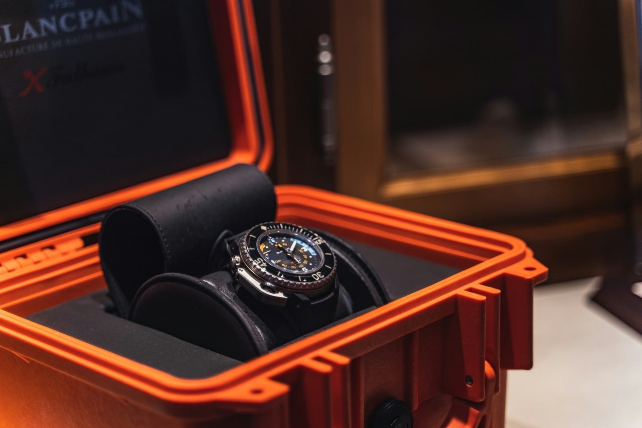 A luxury watch with a black and silver design is displayed in an open vibrant orange case.