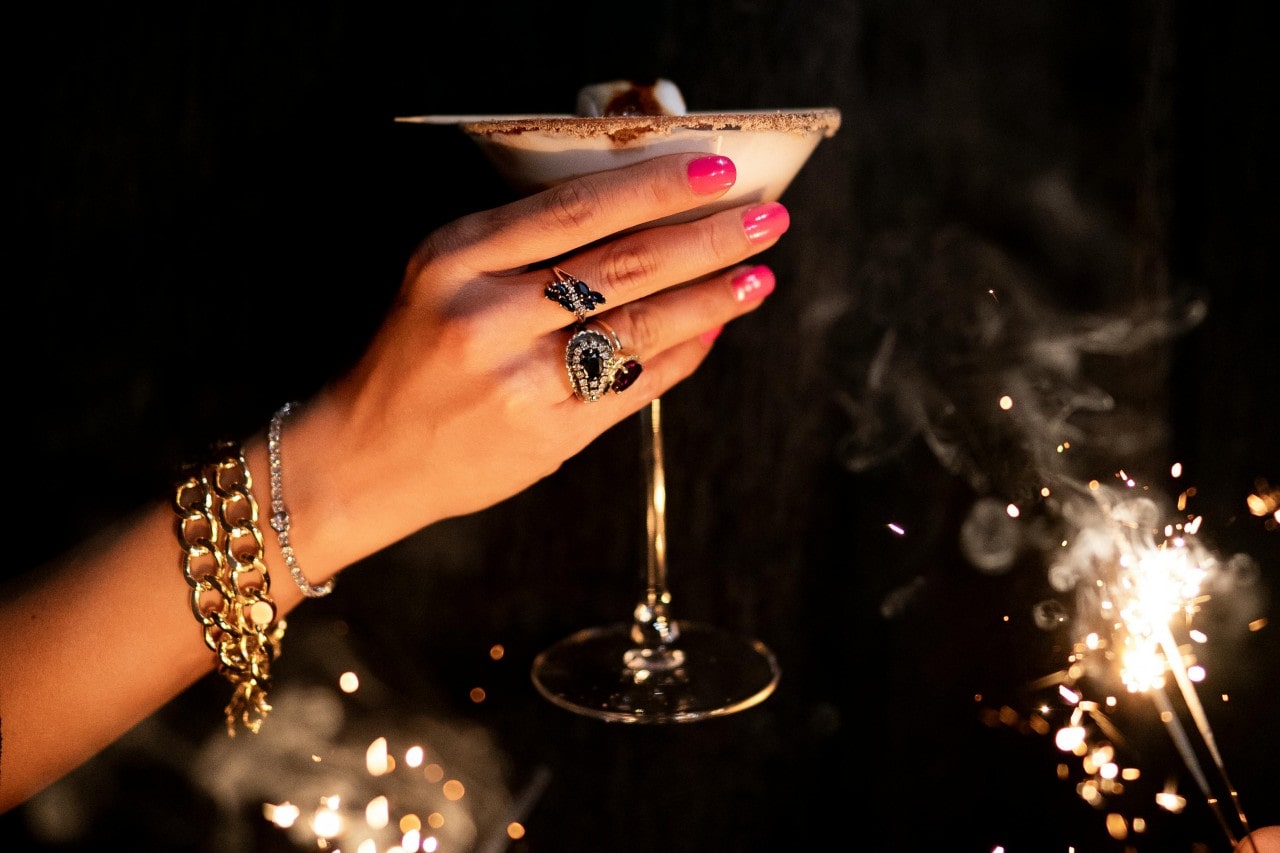 A close-up of a woman’s hand, adorned with eye-catching jewelry, toasting with her martini as sparklers glow in the background.
