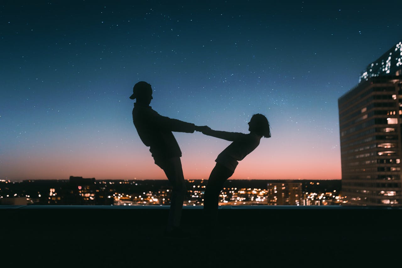 A backlit image of a couple dancing at night on a rooftop with the city skyline for a view.