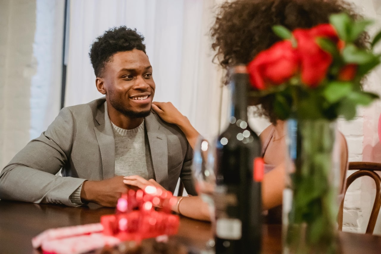 A man and woman holding hands at a wooden table, the woman obscured by a vase of red roses and a bottle of wine, and some gifts.
