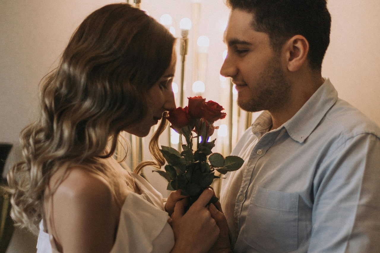 A couple standing closely in a dimly lit room, the woman holding red roses to her nose and admiring their scent.