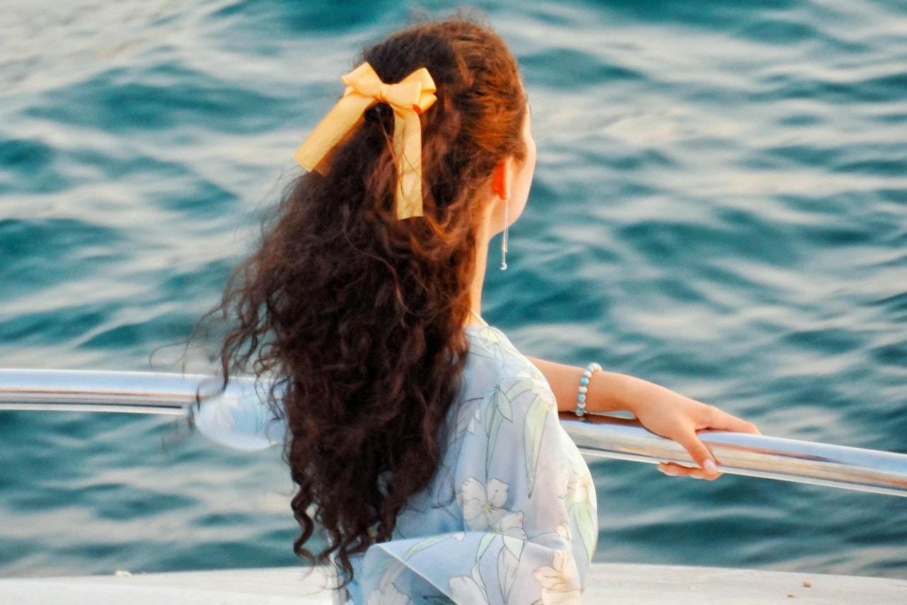 A woman with a yellow bow in her curly hair looking over the edge of a boat rail at the water.