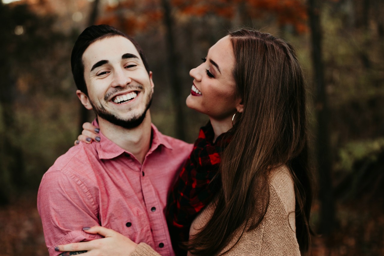 A smiling couple stands in a forest with autumn leaves.