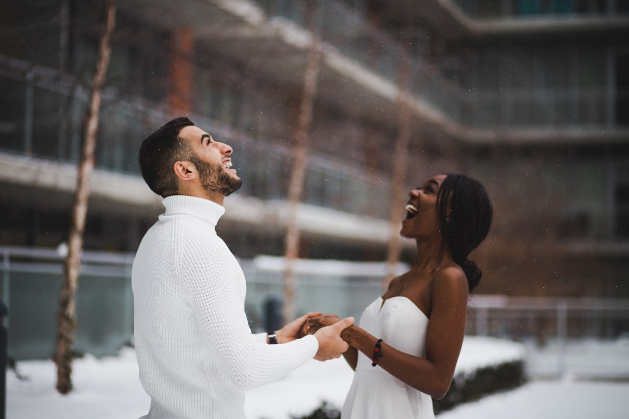 A joyful couple stands in a snowy urban setting, holding hands and looking up, smiling.
