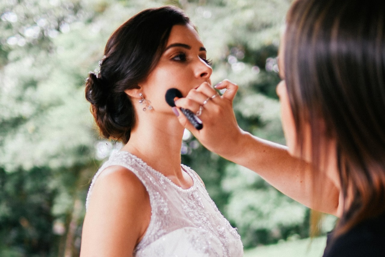 A close-up of a make up artist using a blending brush on a bride’s face while she stands in front of a forestry backdrop.