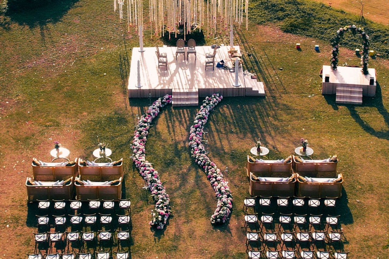 An aerial view of an outdoor wedding venue with a wooden stage, curved flower aisle runners, and guest seating on a lawn.