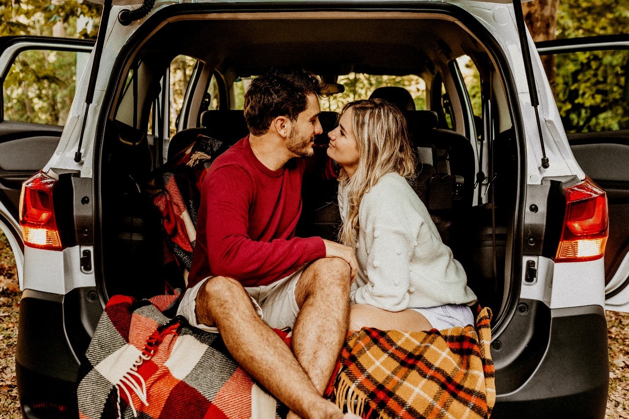 A couple sits on flannel blankets in the trunk of their SUV in a fall forest.