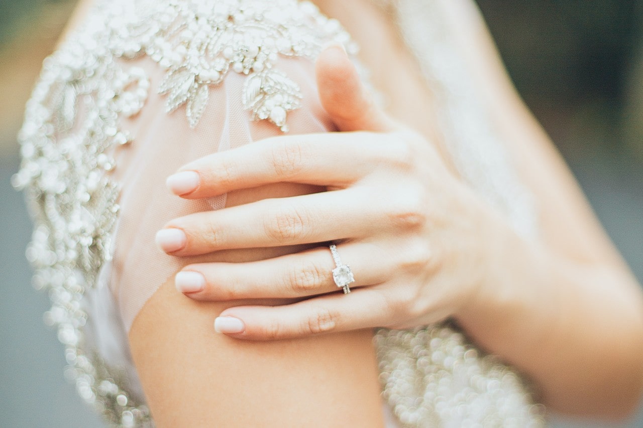a bride’s hand resting on her arm, wearing a white gold cushion cut engagement ring