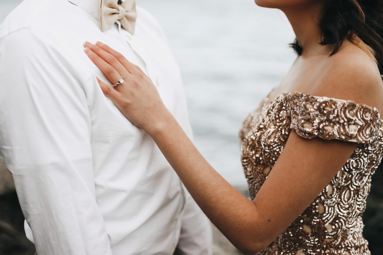 A close-up of a bride in a unique glittery, off-shoulder gold dress gently resting her hand on her groom’s chest, with emphasis on her diamond wedding band.