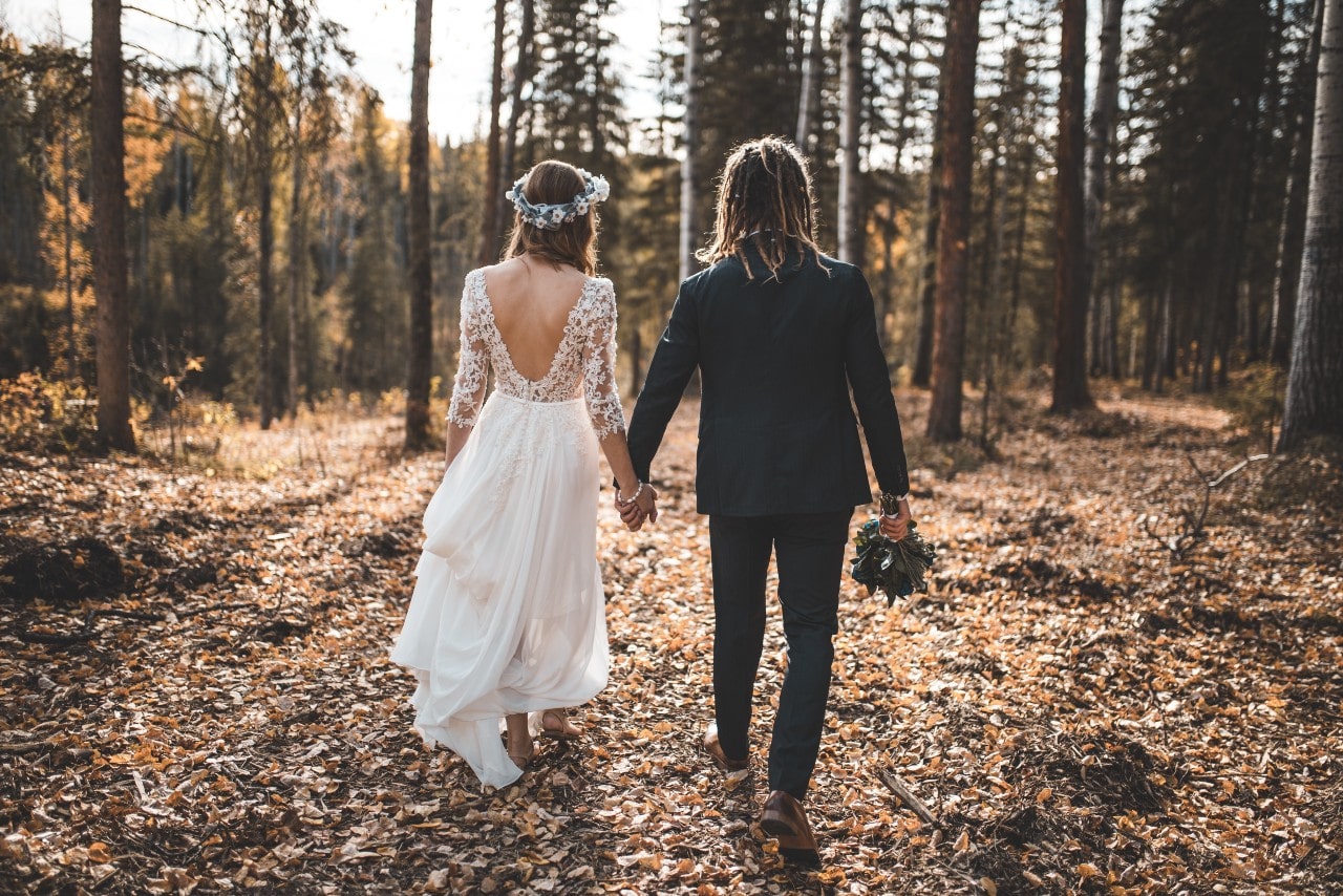 A bride and groom walking hand-in-hand through a sunlit, leafy forest path in autumn.