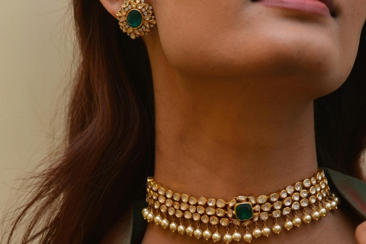 A close-up of a woman wearing a jewelry set consisting of an ornate diamond, pearl, and emerald necklace and matching earrings.