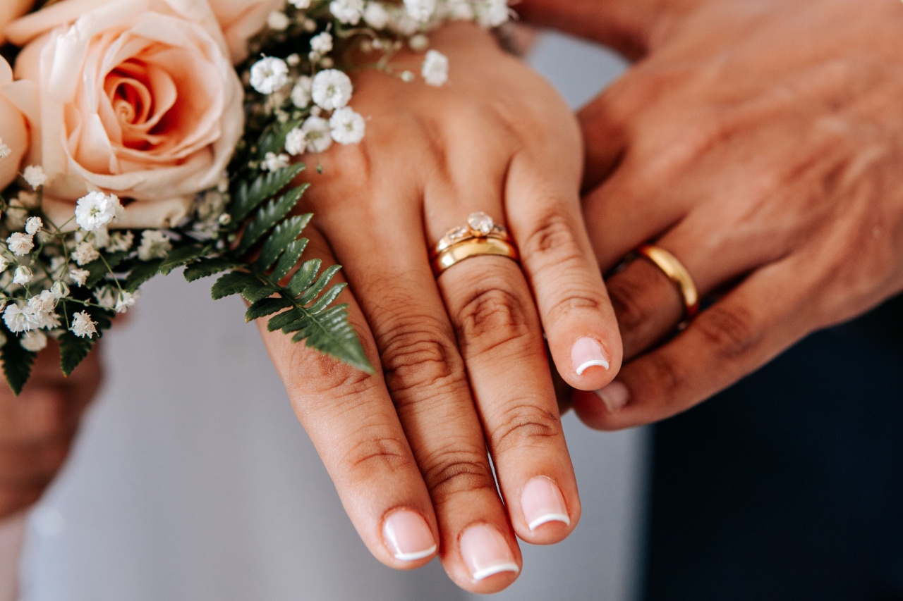 A close-up of a newlywed couple's hands stacked together, showcasing their gold wedding rings next to a pastel pink rose and baby's breath bouquet.