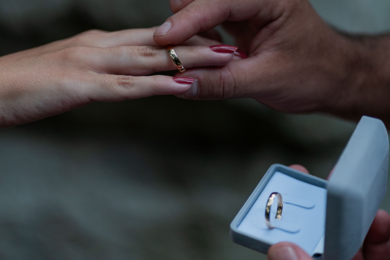 A close-up shot of a hand sliding a gold wedding band onto a finger, while another hand holds open a gray velvet ring box displaying a second matching band.