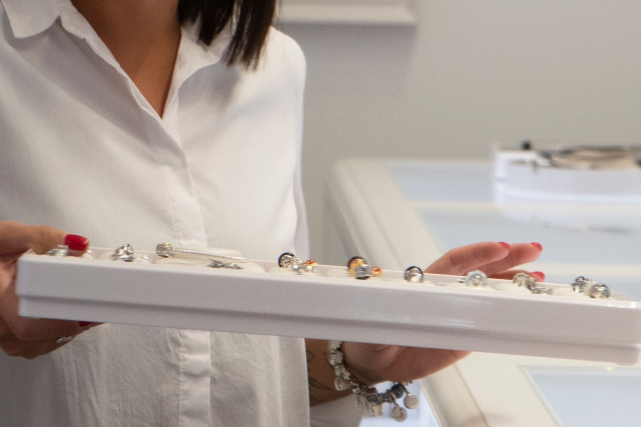 Close-up of a person in a white button-down shirt holding a long tray of engagement rings and charms in a brightly lit jewelry store.