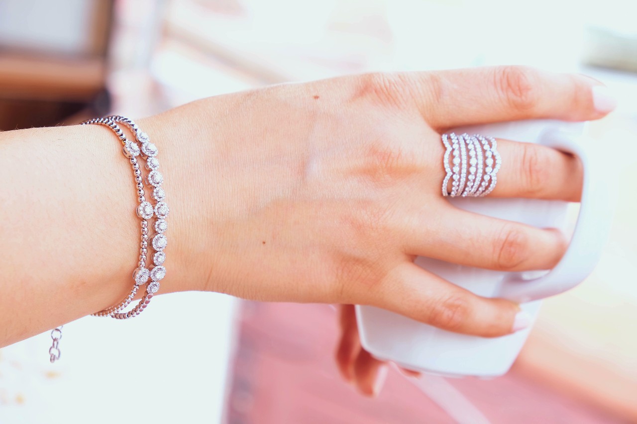 A woman wearing diamond bracelets and fashion rings holds a white mug.