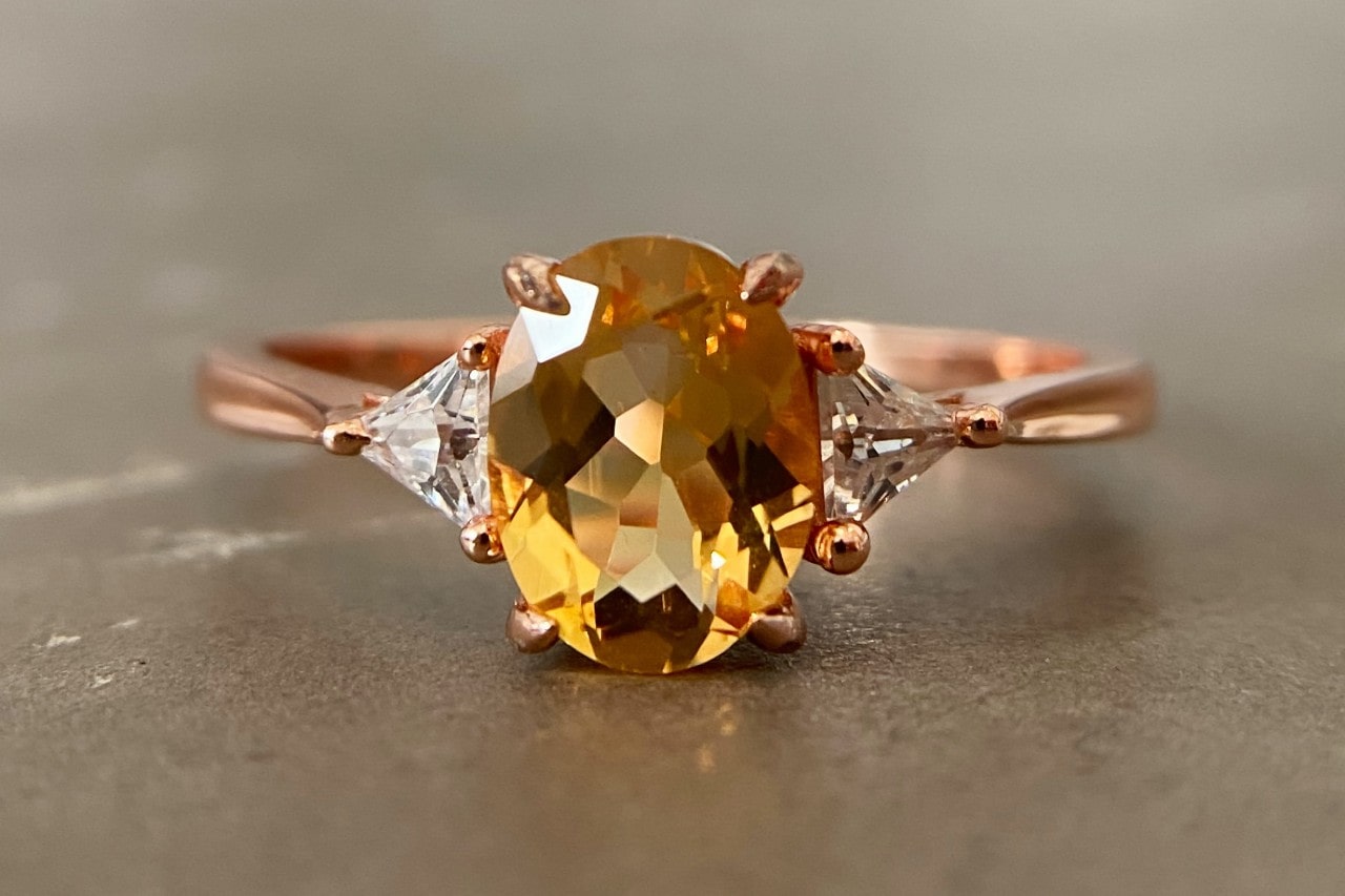 A close-up of a rose gold solitaire ring with an oval cut yellow diamond accented with two smaller triangular diamonds.