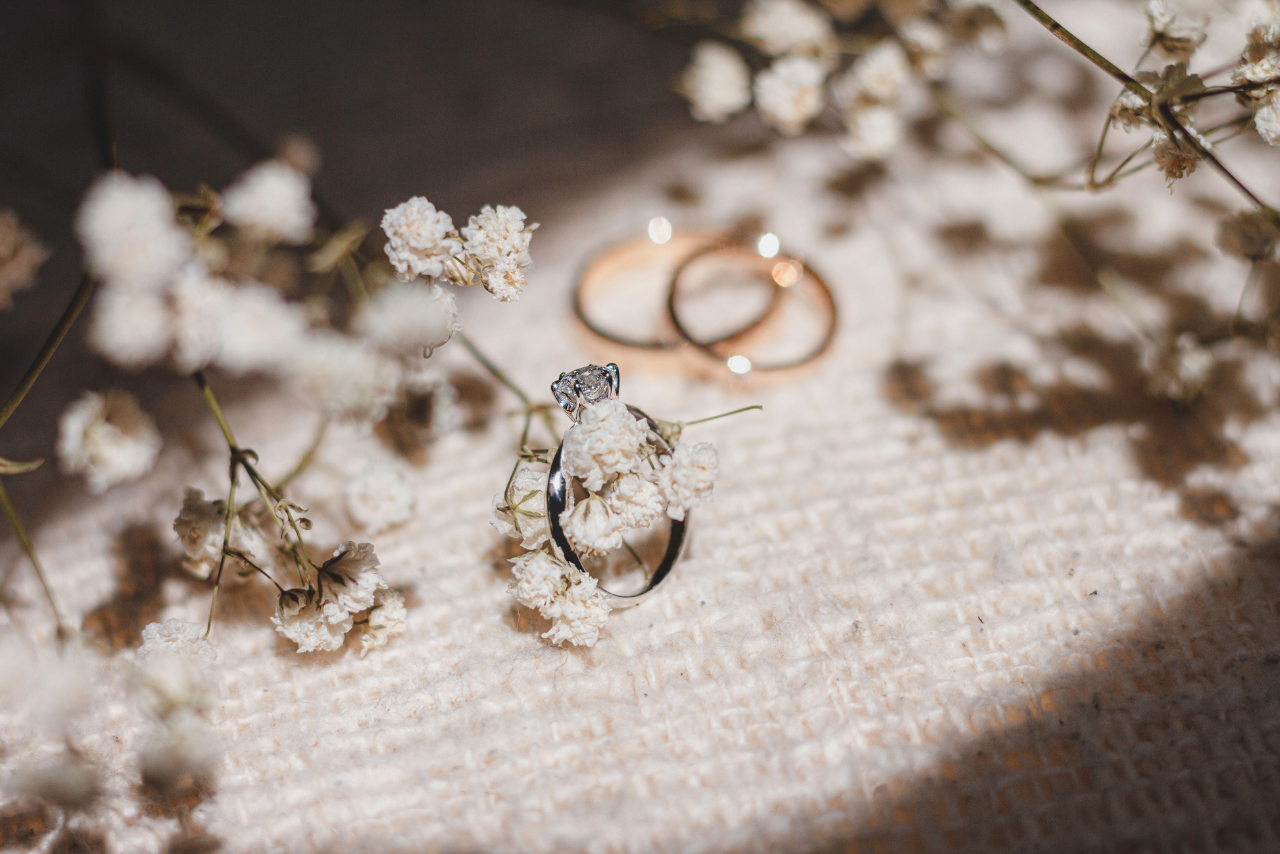 A platinum engagement ring looped around some dried baby’s breath with two yellow gold wedding bands blurred in the background.