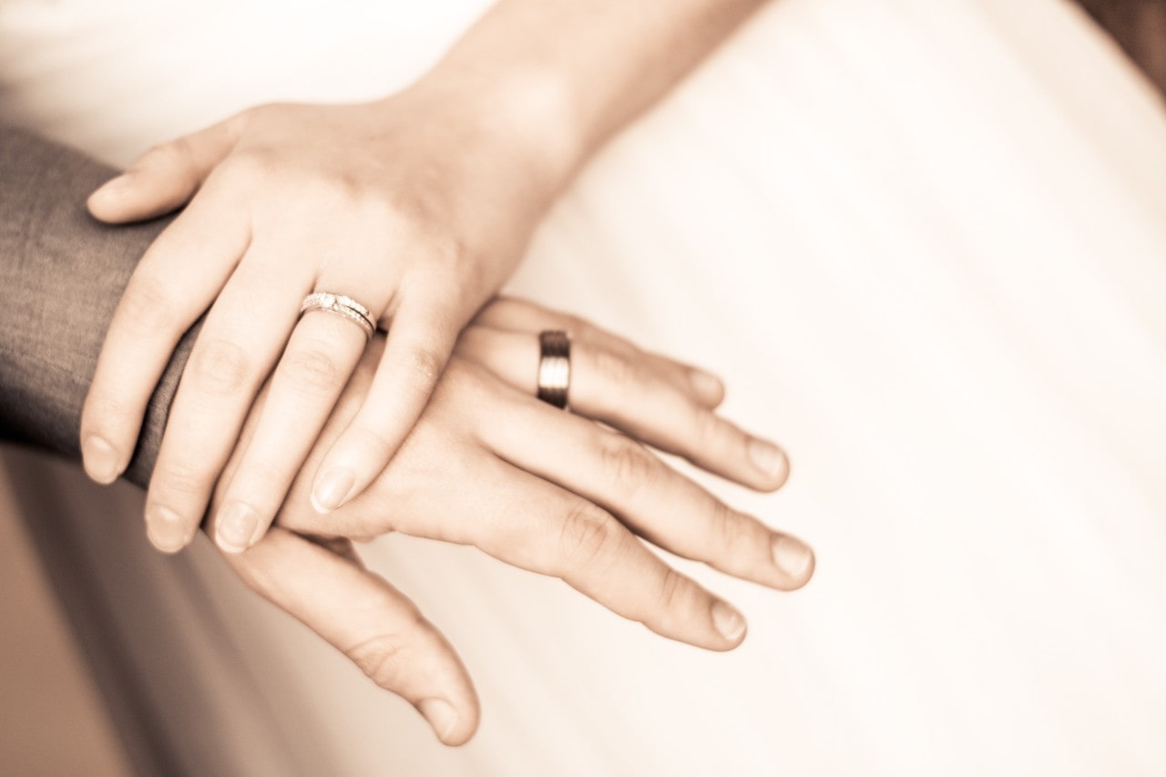 A woman rests her hand on her husband’s, both wearing wedding rings.