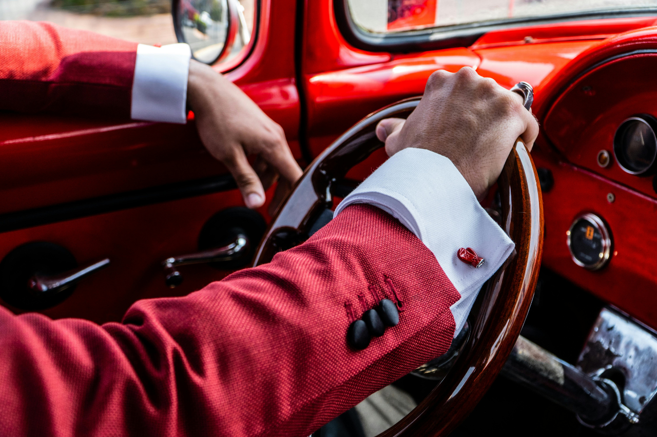 A man in a red suit driving a bright red car, the image focused on his hand on the wheel.