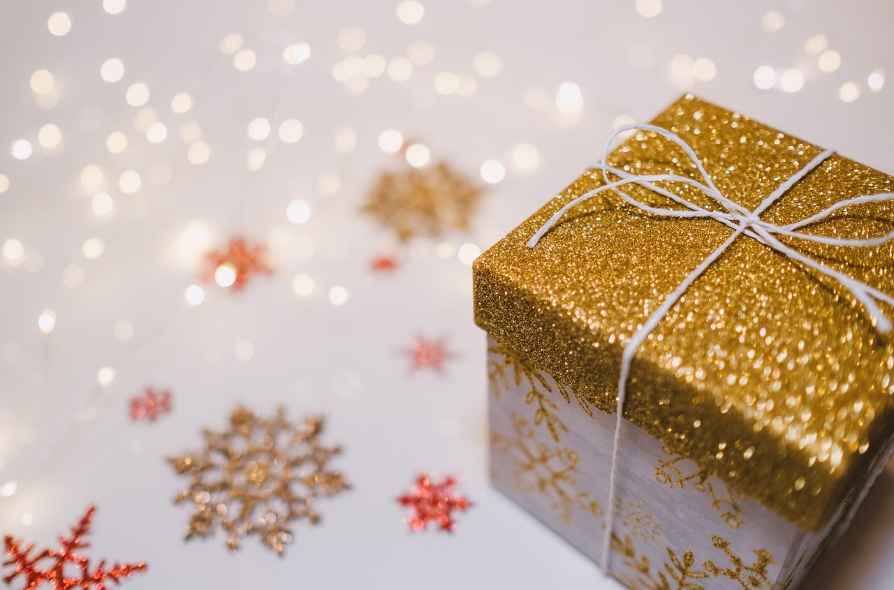 A close-up of a festive gift box with yellow gold glitter top and ribbon in front of glitter snowflakes.