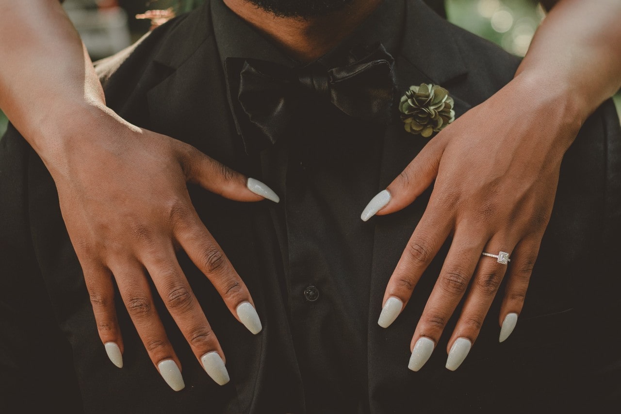 diamond engagement ring on a woman’s hands resting on the chest of her spouse-to-be wearing an all black tux and green flower.