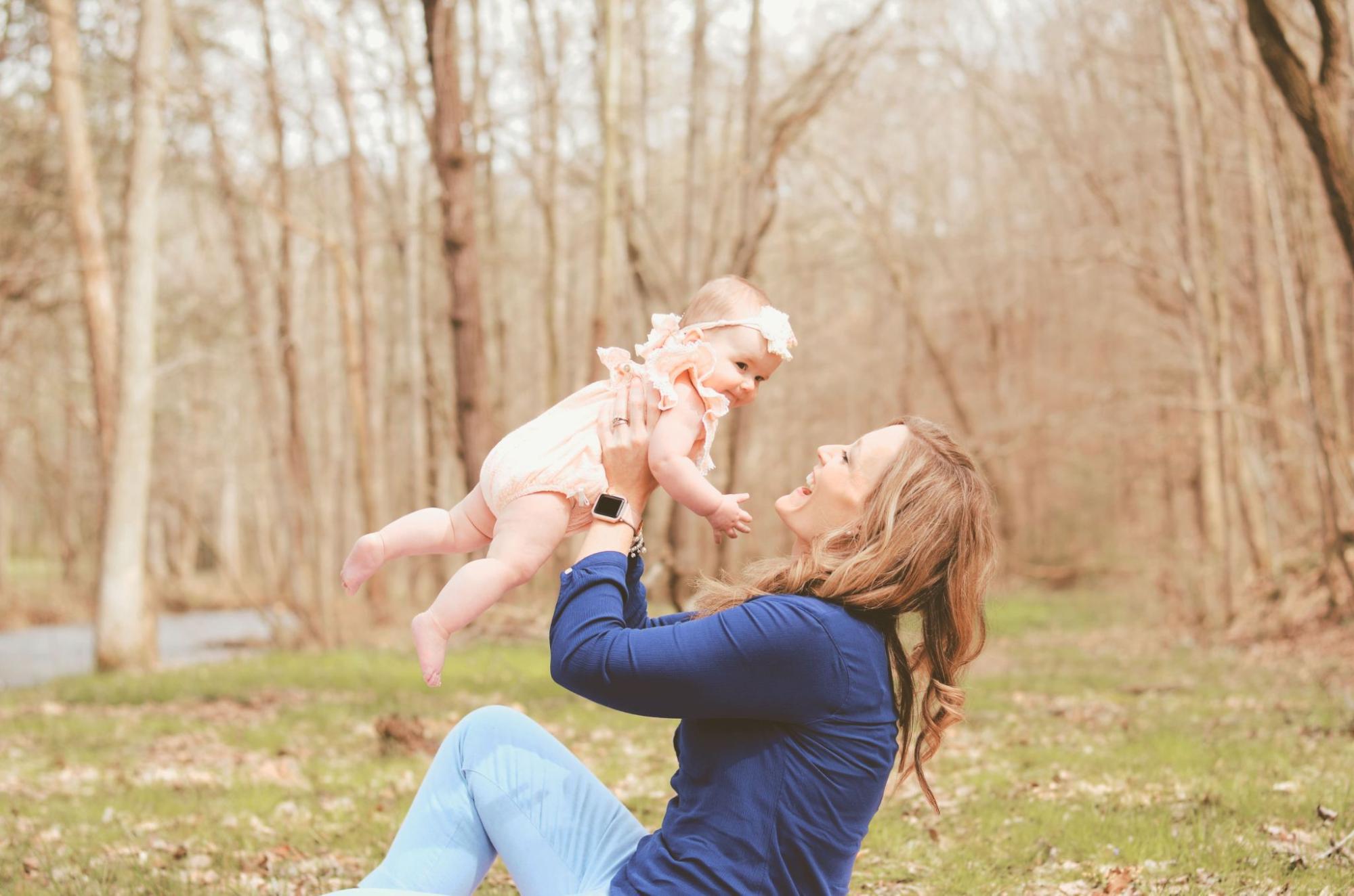 A mother in a blue top joyfully lifts her baby girl, dressed in a peach outfit, while sitting on the grass in a wooded area
