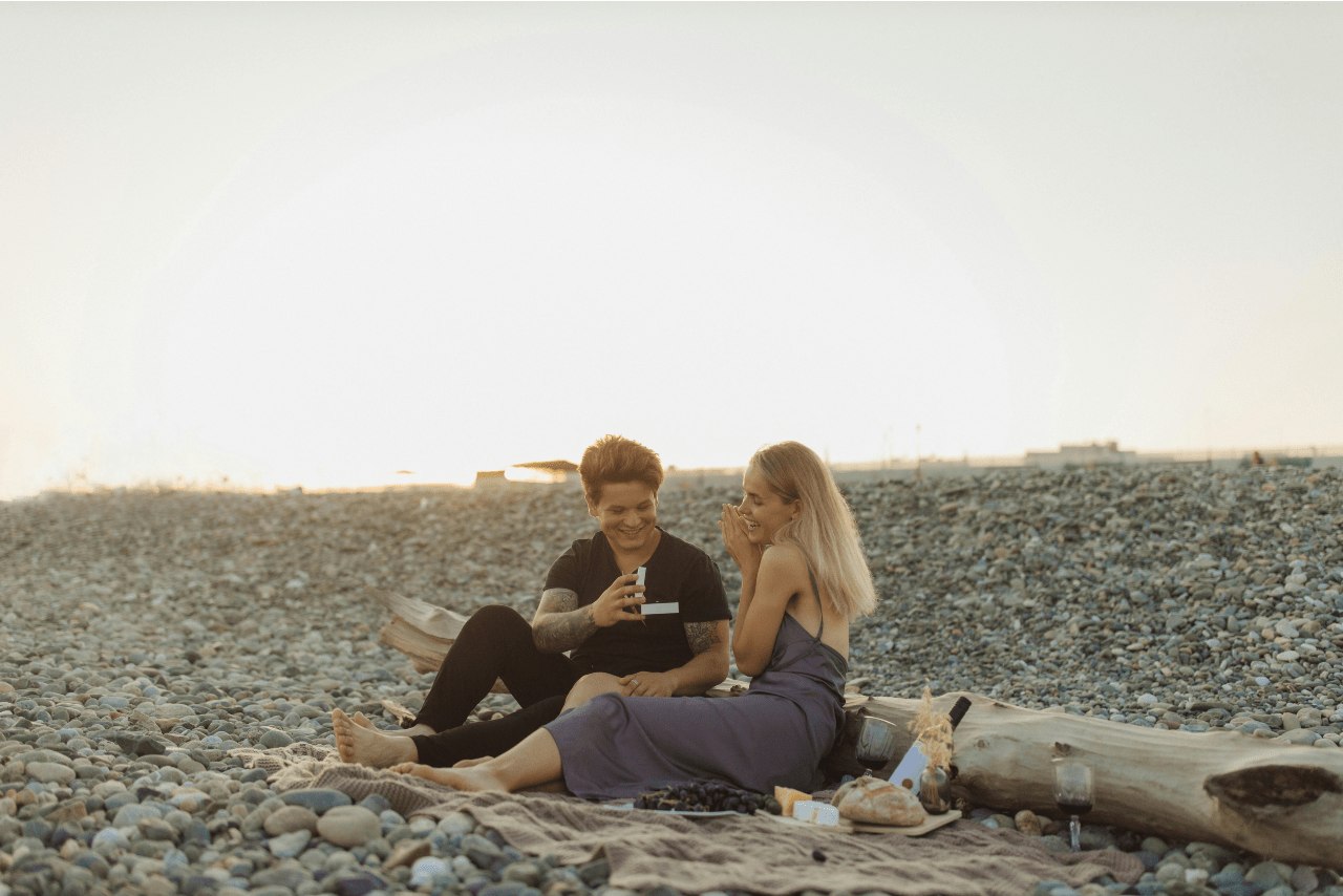 a couple sitting on a rocky beach, one partner proposing to the other with a white ring box