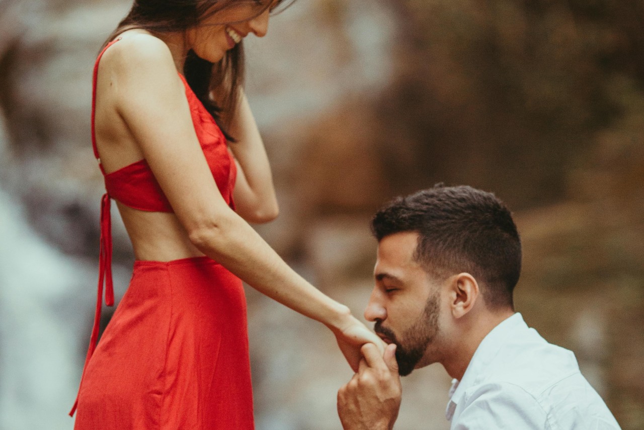 a man kneeling and kissing a woman’s hand who is wearing a red dress