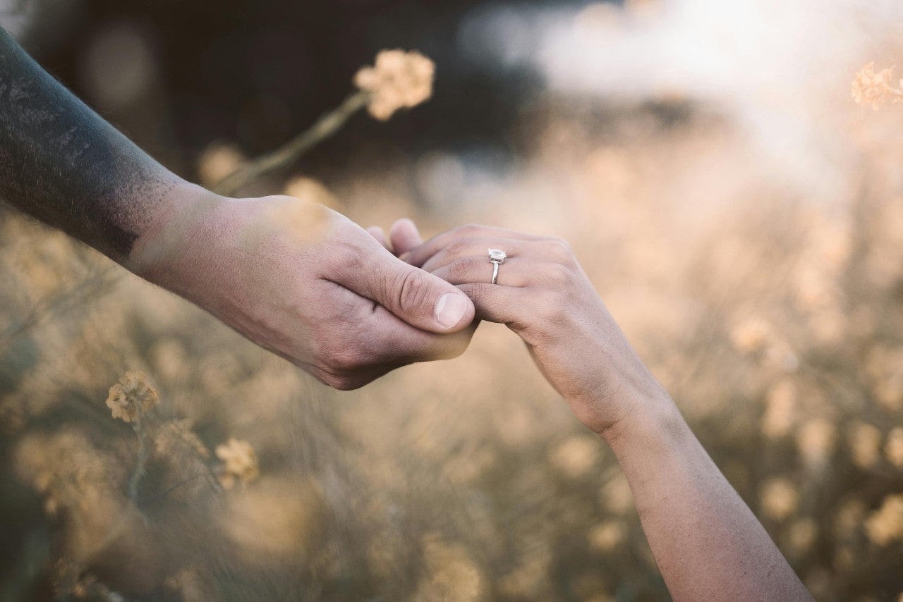 a couple holding hands in a flower field, the woman wearing a silver engagement ring