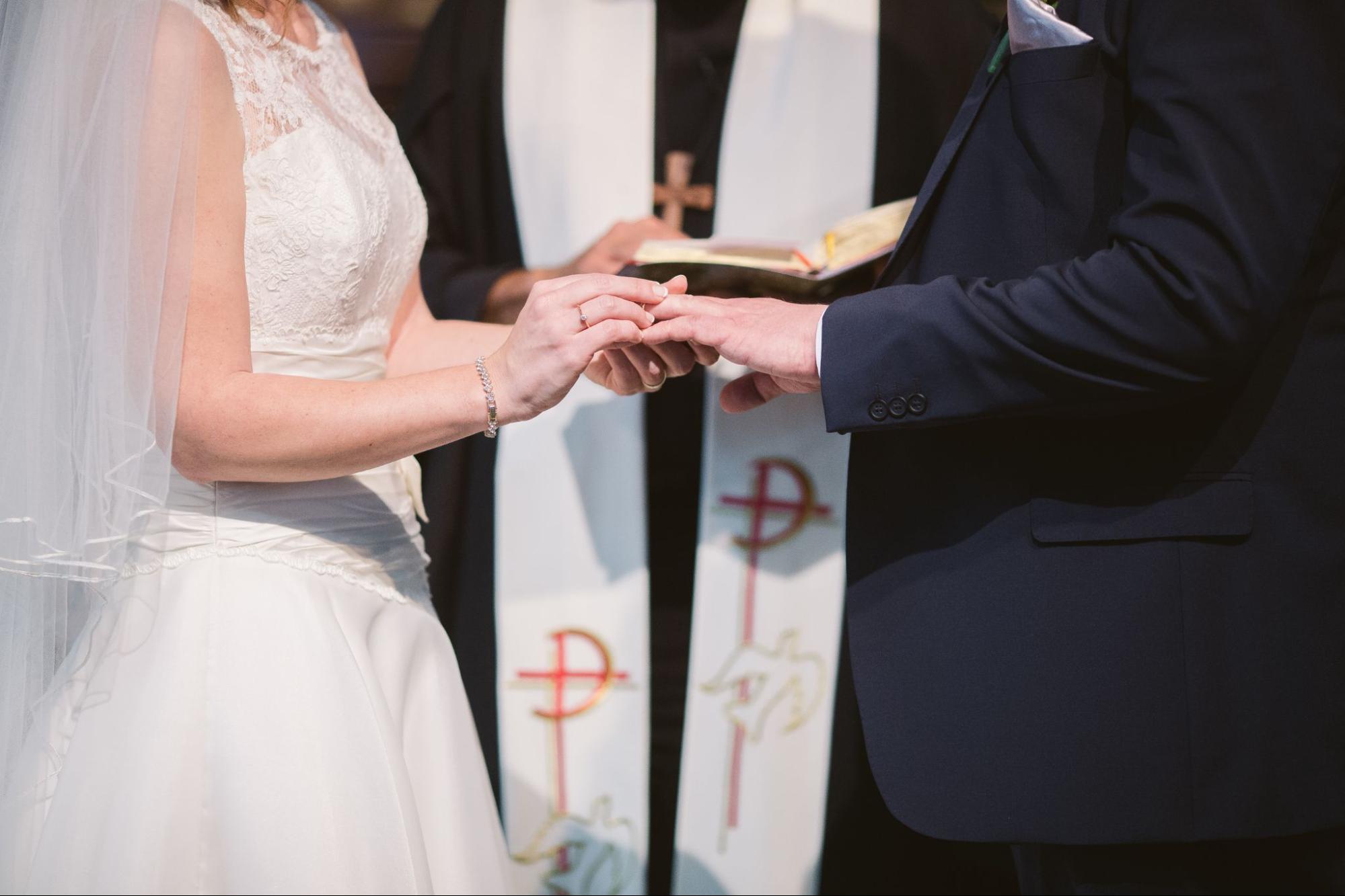 a bride and groom standing in front of a priest, the bride slipping her groom’s ring on his finger