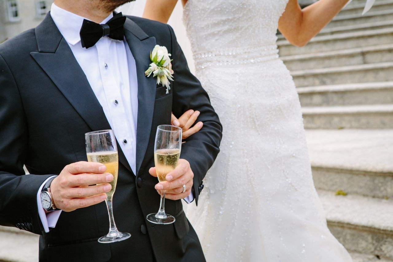 a groom holding two glasses of champagne, his bride holding his arm