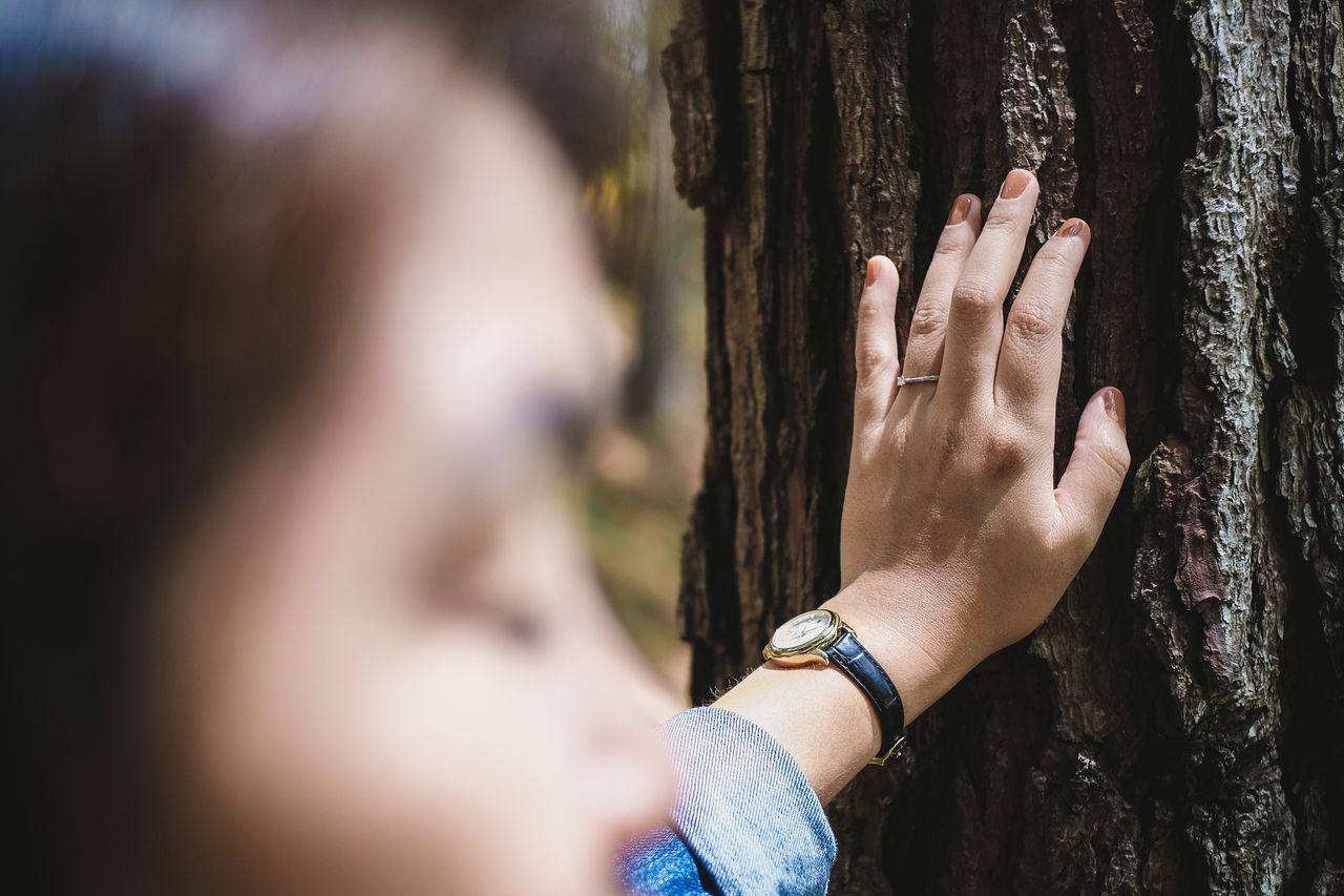 A woman wearing a platinum engagement ring and a watch leans against a tree.