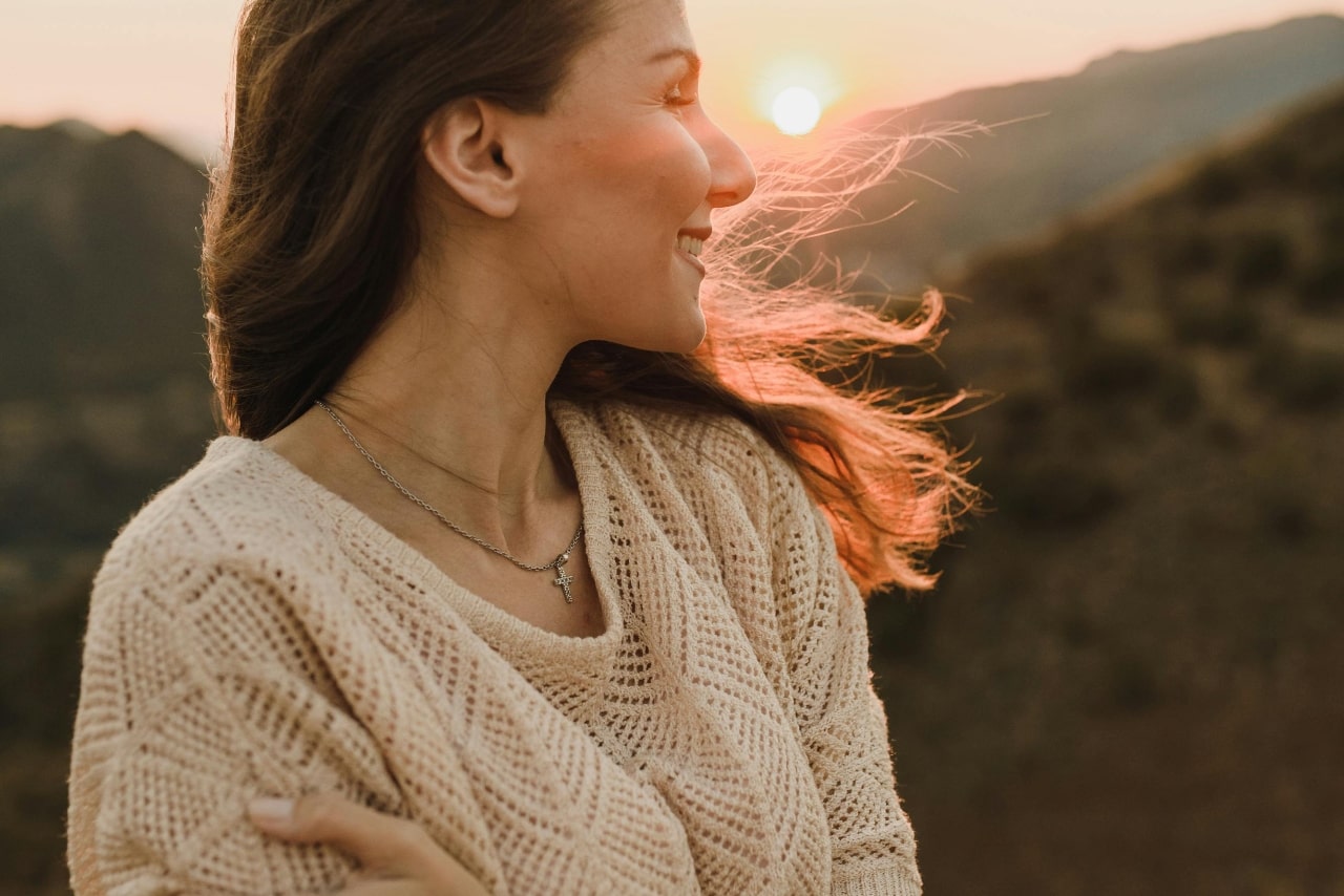 A woman standing in front of the sunlight valley in white knitted sweater with a cross necklace.
