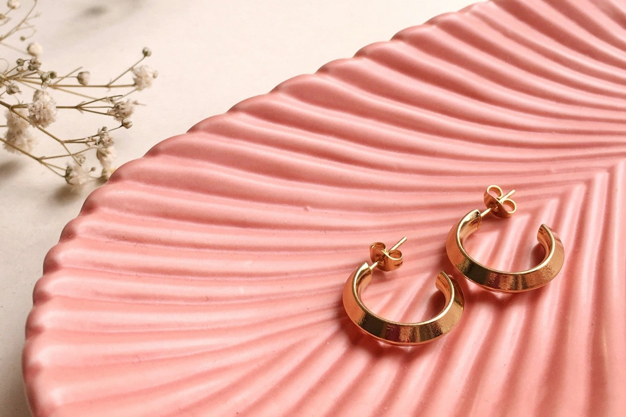 a pair of yellow gold huggie earrings lying in a pink, shell-shaped dish