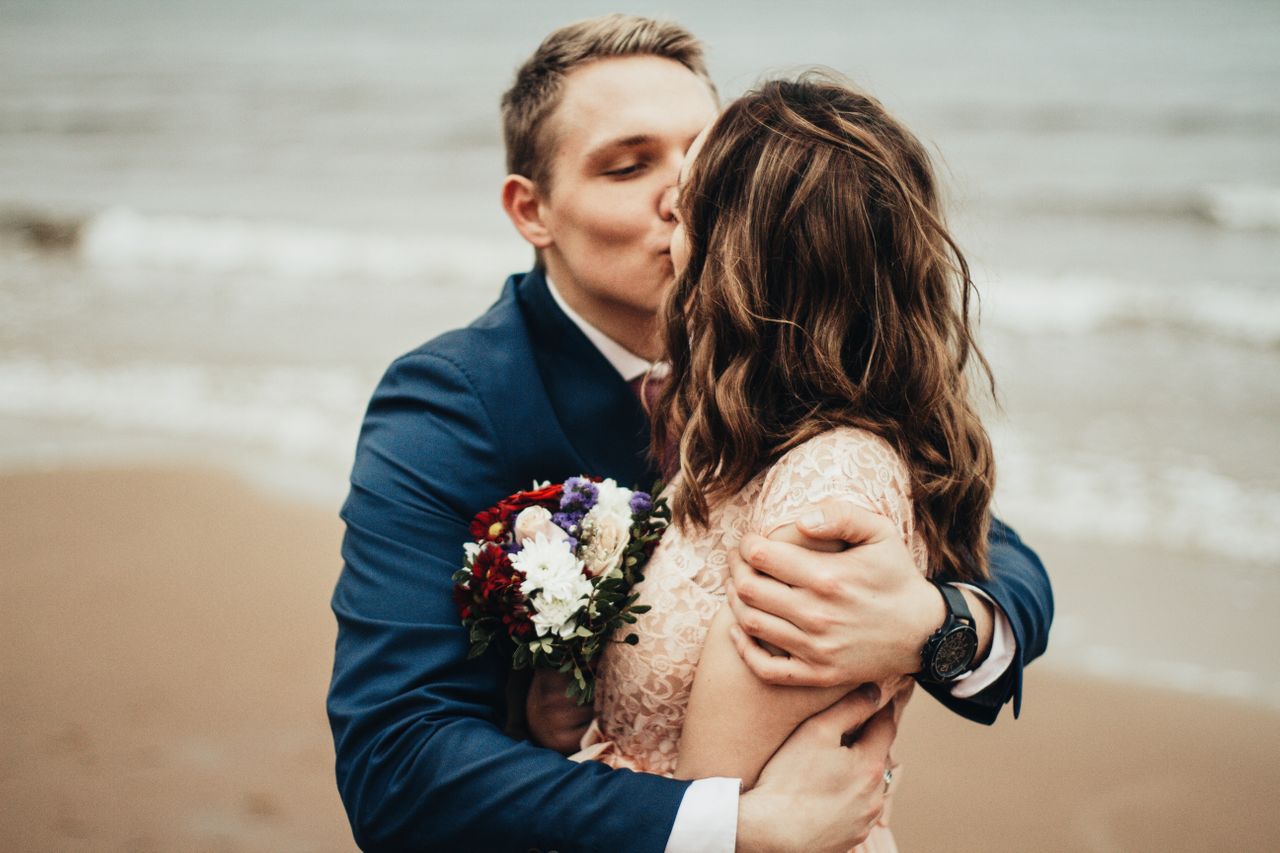 A groom hugs and kisses his bride while on the beach.