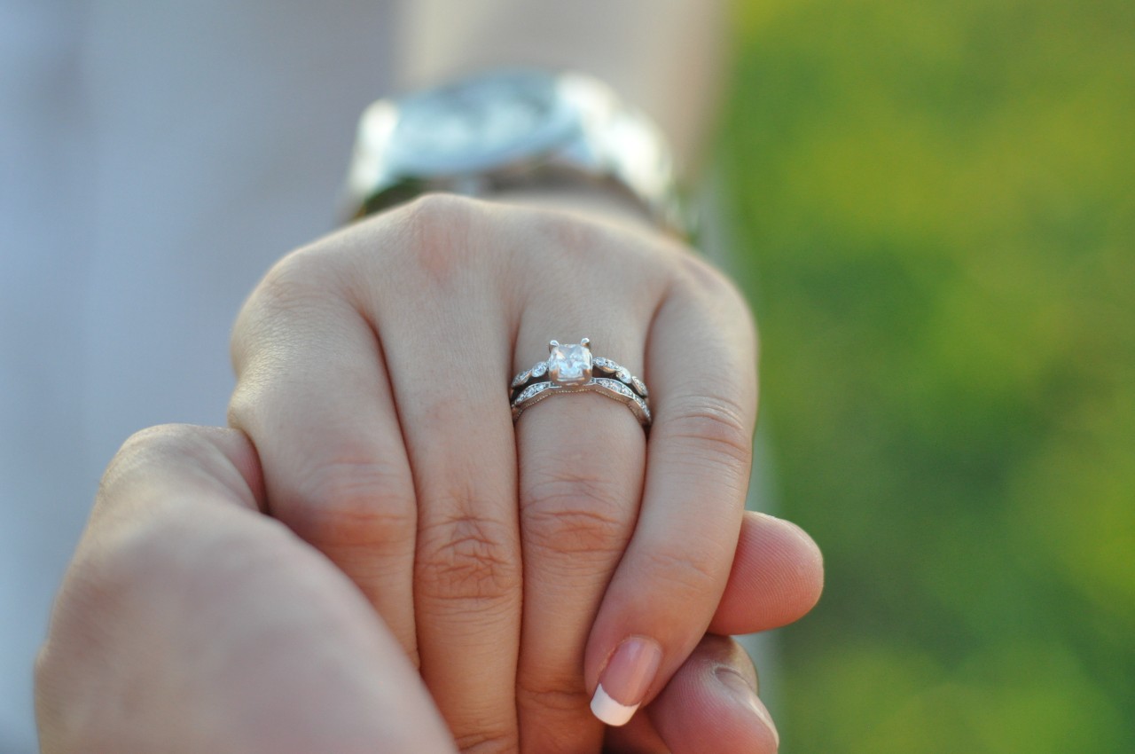 A woman with manicured nails holds her partner’s hand to show off her bridal stack.