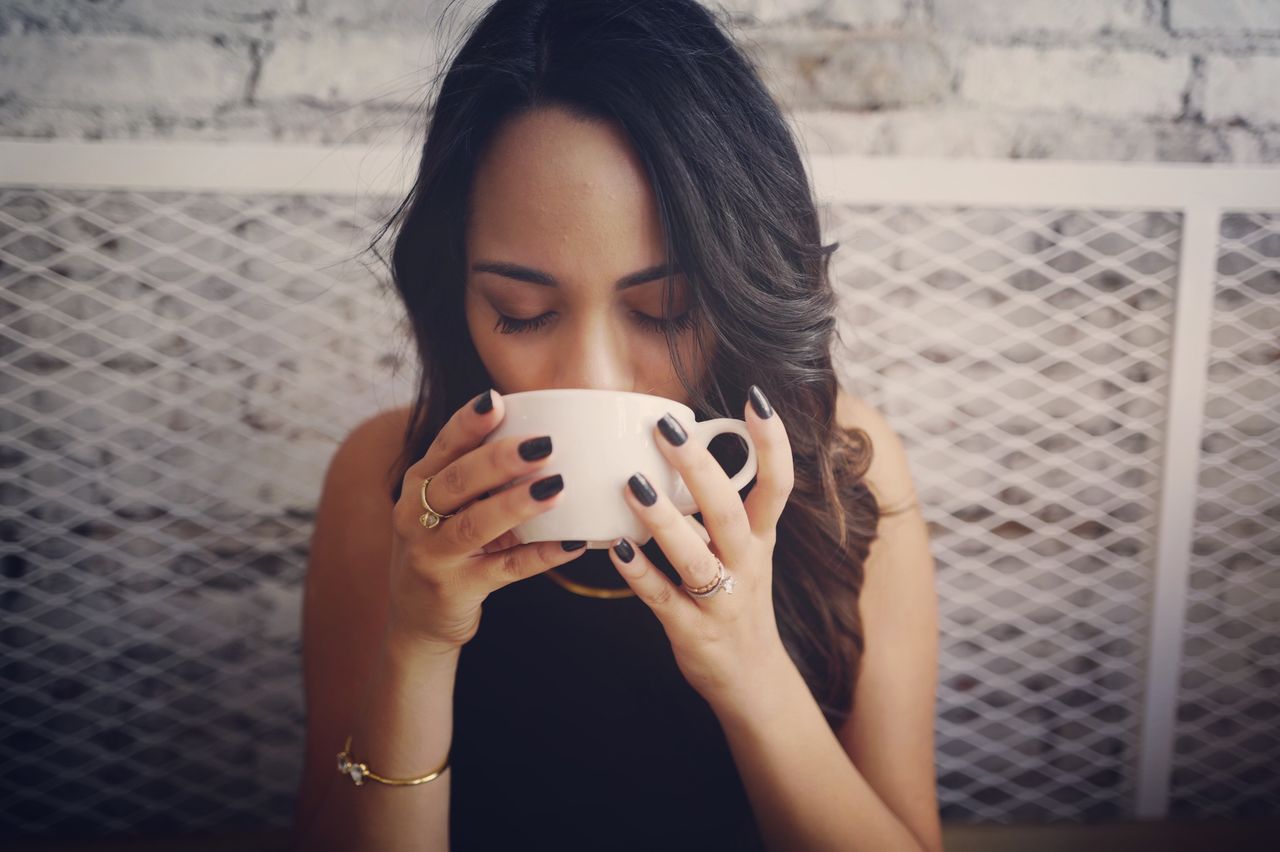 A woman with black nails and gold jewelry, bracelet, necklace, rings, drinking out of a coffee cup