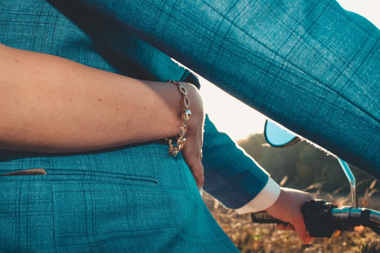 A woman holding onto a man driving a motorcycle. She has a gold bracelet on