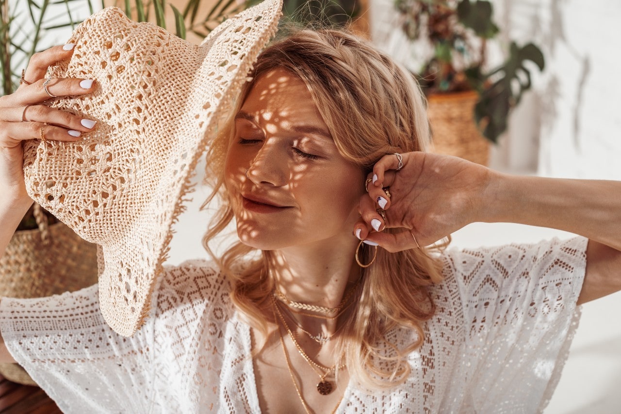 A woman shading herself with a straw hat while she wears multiple pieces of yellow gold jewelry