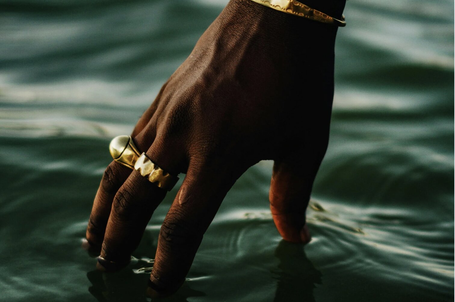 A man wearing yellow gold rings and a yellow gold bracelet dipping his fingers into green water.