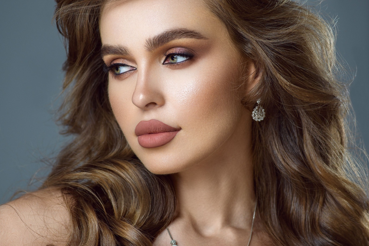 A close-up of a woman with long, wavy brown hair, matte mauve lipstick, and sparkling drop earrings looking to the side.